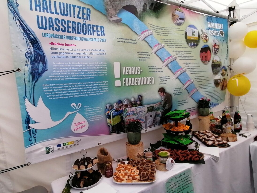 Tisch mit verschiedenen regionalen Produkten, wie Saft und Würsten, vor einer Wand mit dem Thallwitzer Bewerbungsposter »Thallwitzer Wasserdörfer«