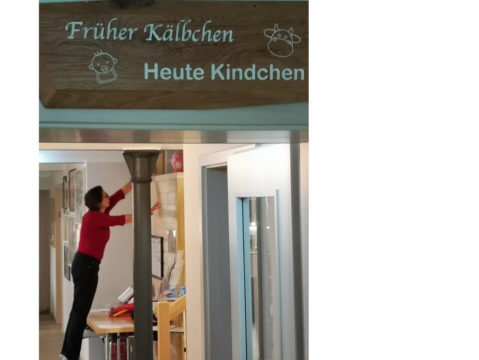 Im Vordergrund steht auf einem Schild über der Tür „Früher Kälbchen, heute Kindchen“, in der Mitte eine alte Säule, dahinter die Hebamme bei der Arbeit