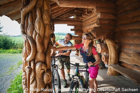 Zwei Fahrradtouristen bestaunen den kettensägegeschnitzten Pfeiler der Hütte in Blockhausen