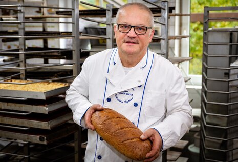 Der Bäckermeister steht mit einem Brot in den Händen in der Backstube.