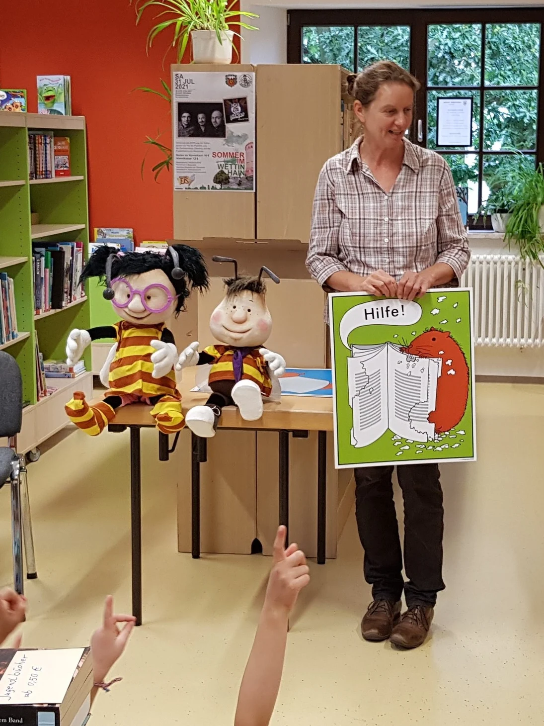 Lesebienchen aus Stoff sitzen auf einem Tisch neben der stehenden Projektkoordinatorin in der Bibliothek