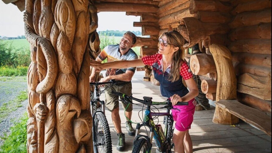 Zwei Fahrradtouristen bestaunen den kettensägegeschnitzten Pfeiler der Hütte in Blockhausen