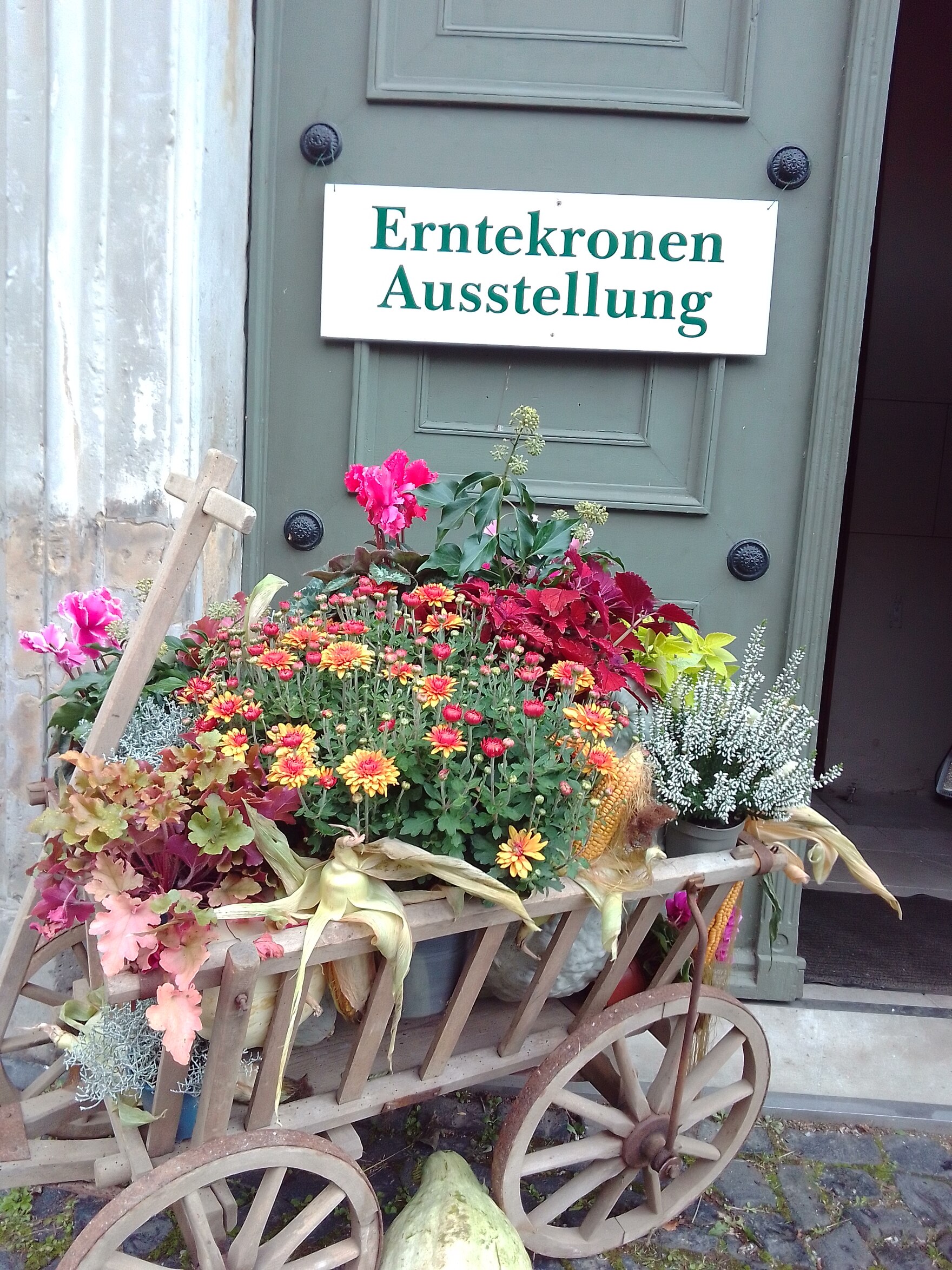 Vor der Tür kleiner Leiterwagen auf Holz bepackt mit Herbstblumen in Töpfen, an der grauen Tür ein Schild "Erntekronen Ausstellung"