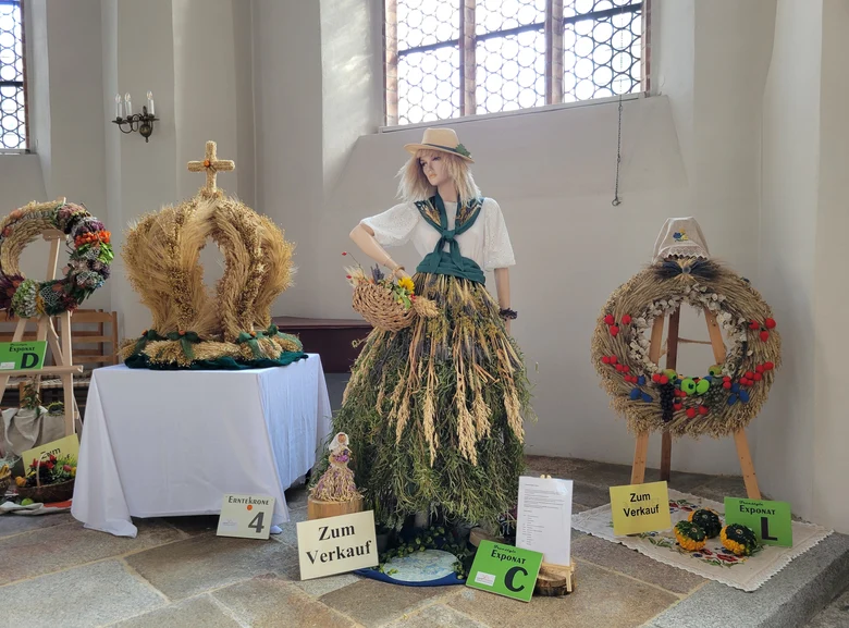 verschiedene Exponate der Ausstellung 2025 mit gebundenen Kränzen und Kronen sowie einer Puppe mit Kleidung aus Naturmaterial als Freestyle Exponat