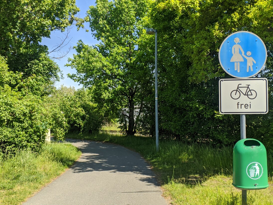 Blick auf Weg, umsäumt mit Bäumen, blaues Verkehrsschild: Fußgängerweg und weißes Schild Fahrräder frei im Vordergrund, darunter ein Mülleimer