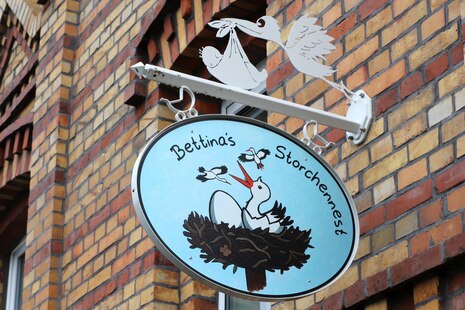 Ovales Schild mit Storchenmotiv und Aufschrift Bettinas Storchennest, angebracht an einem Halter an der Hausfassade
