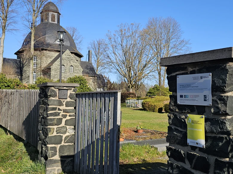 Friedhofseingang mit steinernen Torpfeilern, an dem Schilder befestigt sind, im Hintergrund der Friedhof und die Lutherkirche
