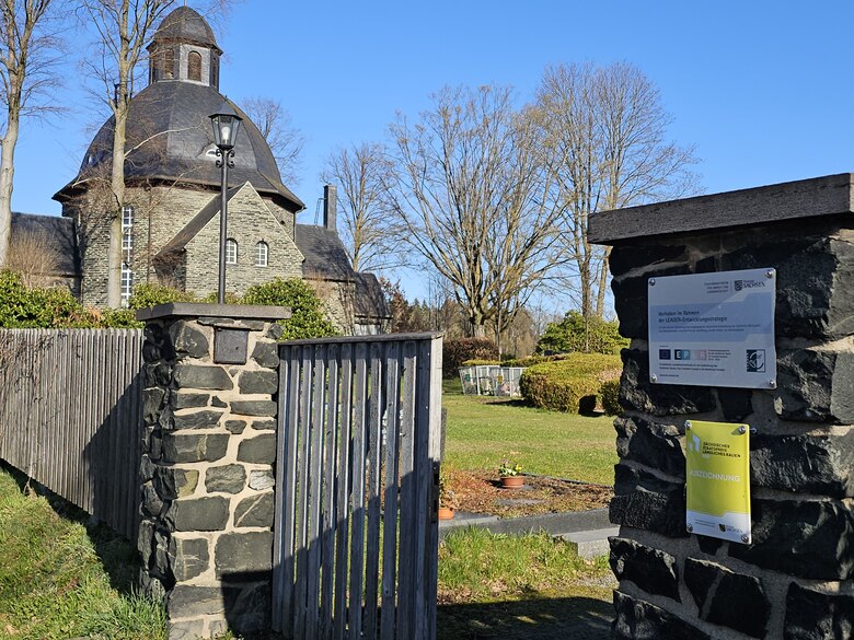 Friedhofseingang mit steinernen Torpfeilern, an dem Schilder befestigt sind, im Hintergrund der Friedhof und die Lutherkirche