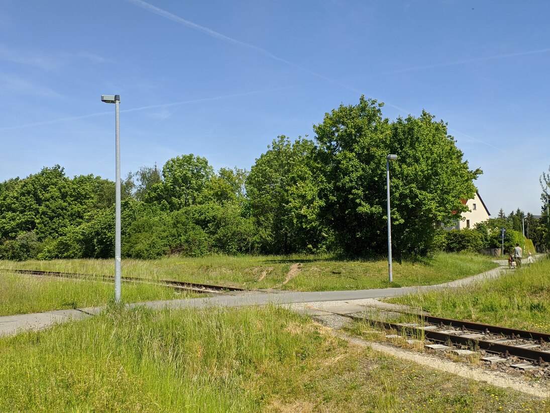 Bahngleise bei Querungsstelle mit Asphalt überzogen, flankiert von Straßenlaternen, Tagesmutter im Hintergrund