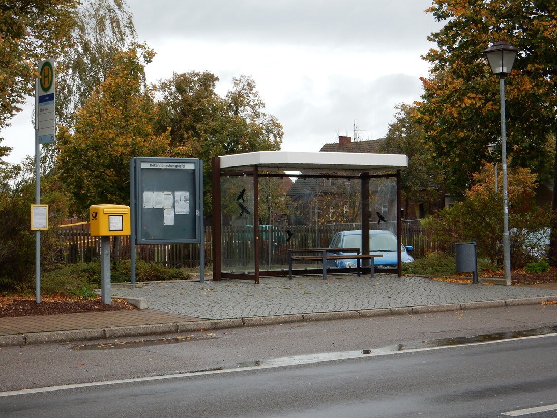 Bushaltestelle mit neuem transparent gestaltetem Fahrgastunterstand, links daneben eine Bekanntmachungstafel und Postbriefkasten
