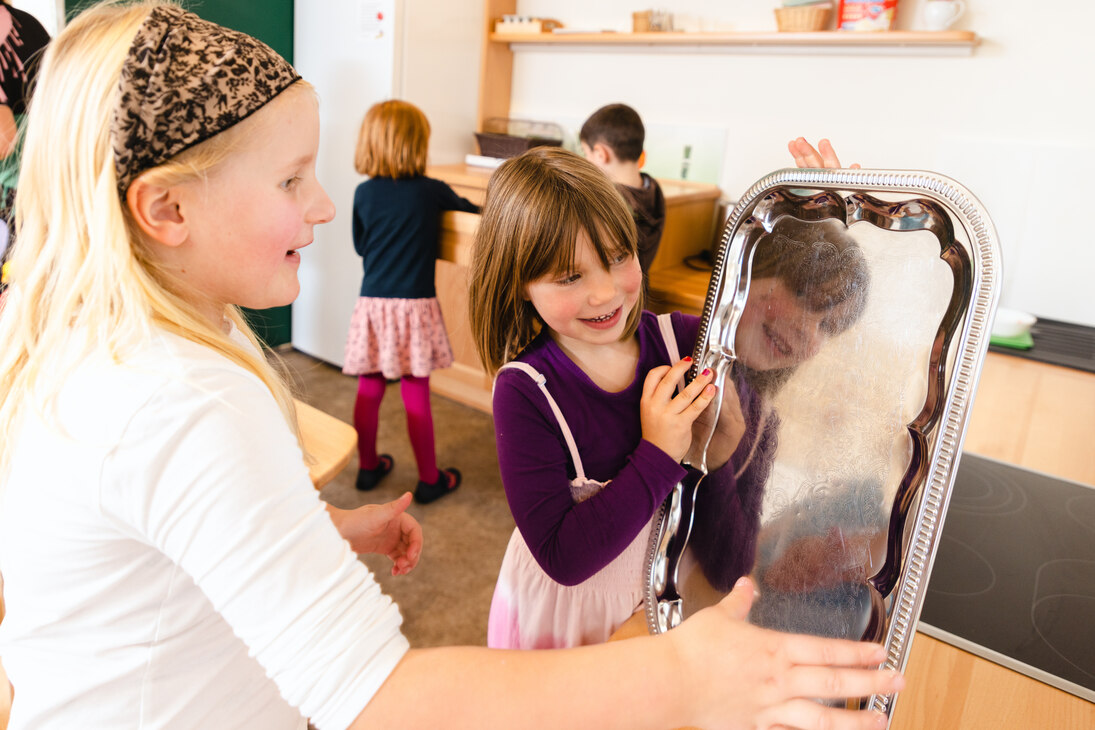 Im Hintergrund spielende Kinder im umgebauten Speiseraum mit eingebauter Kinderküche im Gebäude der Kindertagesstätte Kinder-Lehm-Haus in Grimma. Im Vordergrund sieht ein Mädchen ihr Spiegelbild in einem silbernen Tablett.