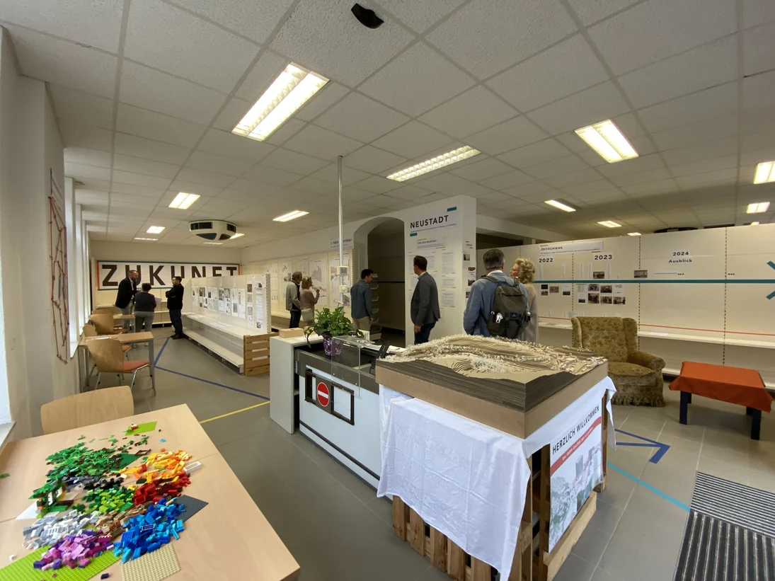 In einem modernen Ausstellungsraum betrachten mehrere Personen verschiedene Präsentatio-nen zu Architektur und Stadtplanung. Auf einem Tisch liegen bunte LEGO-Steine, und ein großes Modell sowie Infotafeln zeigen Zukunftsprojekte unter dem Motto „ZUKUNFT“