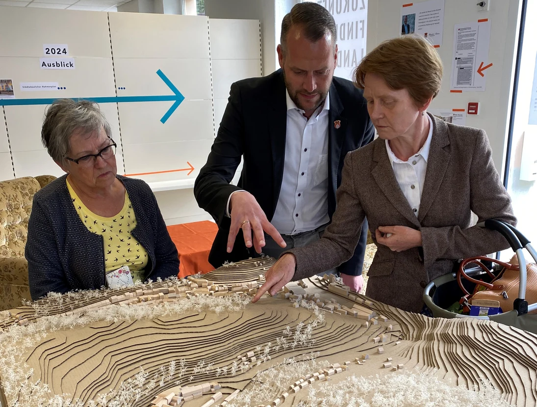 Drei Personen betrachten gemeinsam ein detailliertes Landschaftsmodell mit Hügeln, Gebäu-den und Vegetation. Eine Person zeigt auf einen Bereich des Modells, während die anderen aufmerksam zuhören und diskutieren.