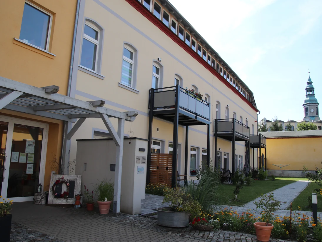 zwei-etagige Gebäude mit Terrasse und Balkonen, im Hintergrund ist der Kirchturm der Niederoderwitzer Kirche zu sehen