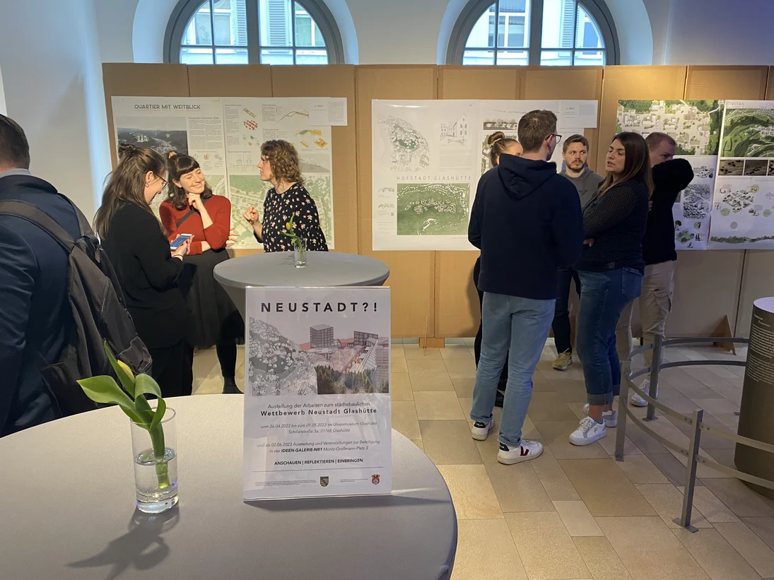 Die Aufnahme zeigt eine Ausstellung in einem hellen Raum mit großen Fenstern, bei der mehrere Personen Architekturprojekte auf Präsentationstafeln betrachten. Im Vordergrund steht ein kleiner Tisch mit einem Flyer und einer Vase mit einer grünen Pflanze.