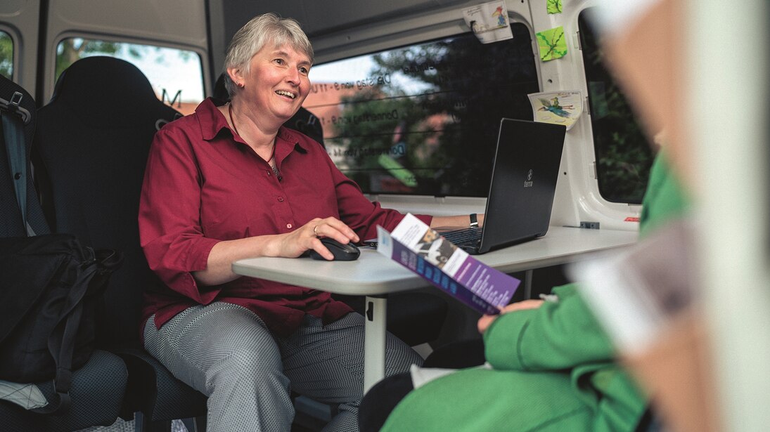 Die Diplomsozialarbeiterin sitzt im umgebaute Kleinbus vor ihrem Laptop und berät eine Hilfesuchende, die Informationsmaterial in der Hand hält.