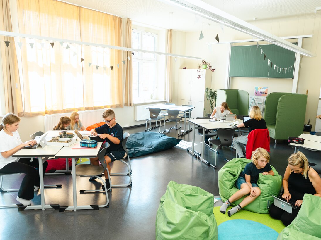 Blick in das mit Wimpeln geschmückt Klassenzimmer. Kinder arbeiten an gruppierten Tischen oder auf bunten Sitzsäcken sitzend. Alle Kinder haben Tablets. Die Lehrerin erklärt einem Jungen etwas.