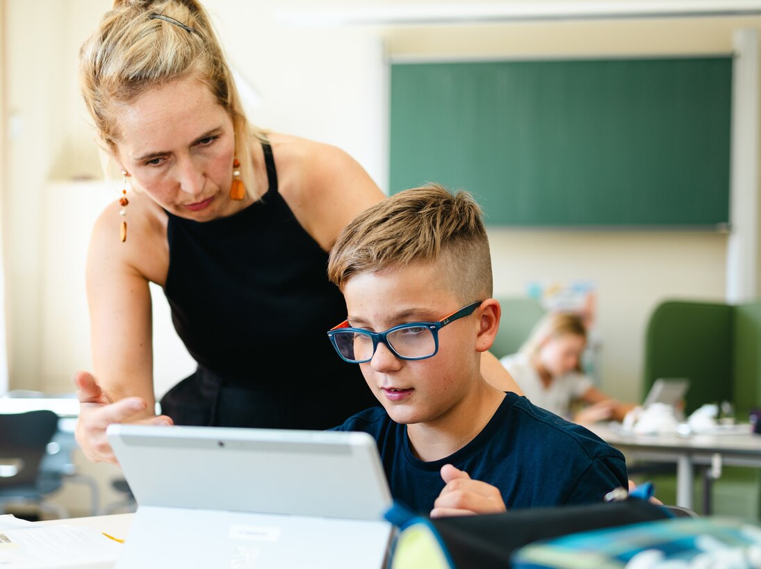 Eine Lehrerin des Freien Gymnaisums Borsdorf beugt sich zu einem Schüler, um ihm etwas zu erklären. Der Junge sitzt mit einem Tablet an einem Tisch im Klassenzimmer.