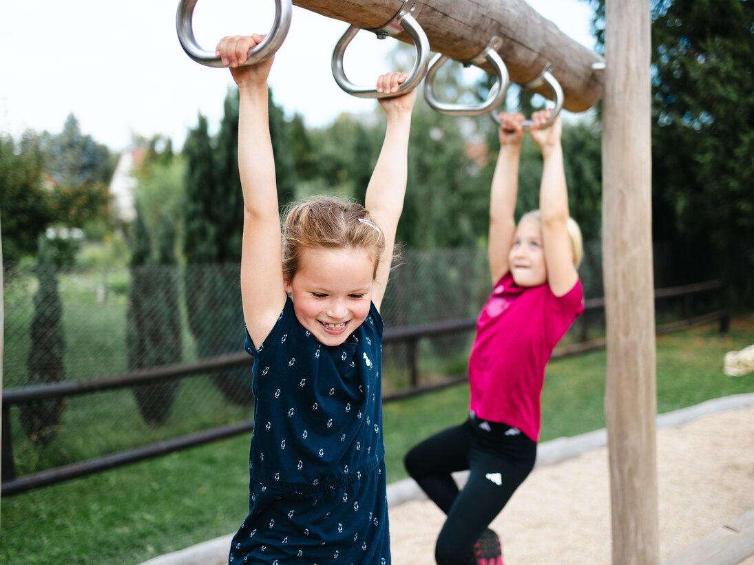 Zwei Mädchen hangeln sich an Griffen an einem Holzbalken auf dem Spielplatz entlang.