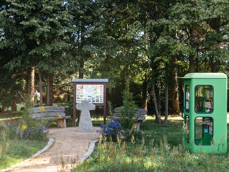 Ein kleiner Weg führt über eine Wiese zum Treffpunkt mit Holzbänken, einer Infotafel und einem Steinkreuz in der Mitte. Im Hintergrund Bäume.