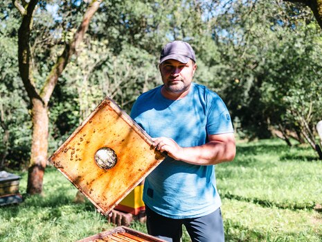 Ein Mann hält einen Holzrahmen gefüllt mit Honigwaben in den Händen. Im Hintergrund der Imkergarten Rathmannsdorf.