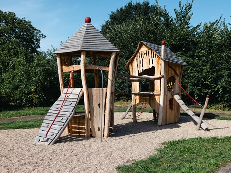 Zwei Kletter- und Spielhäuser aus Holz auf dem Spielplatz, verbunden durch eine Hängebrücke.