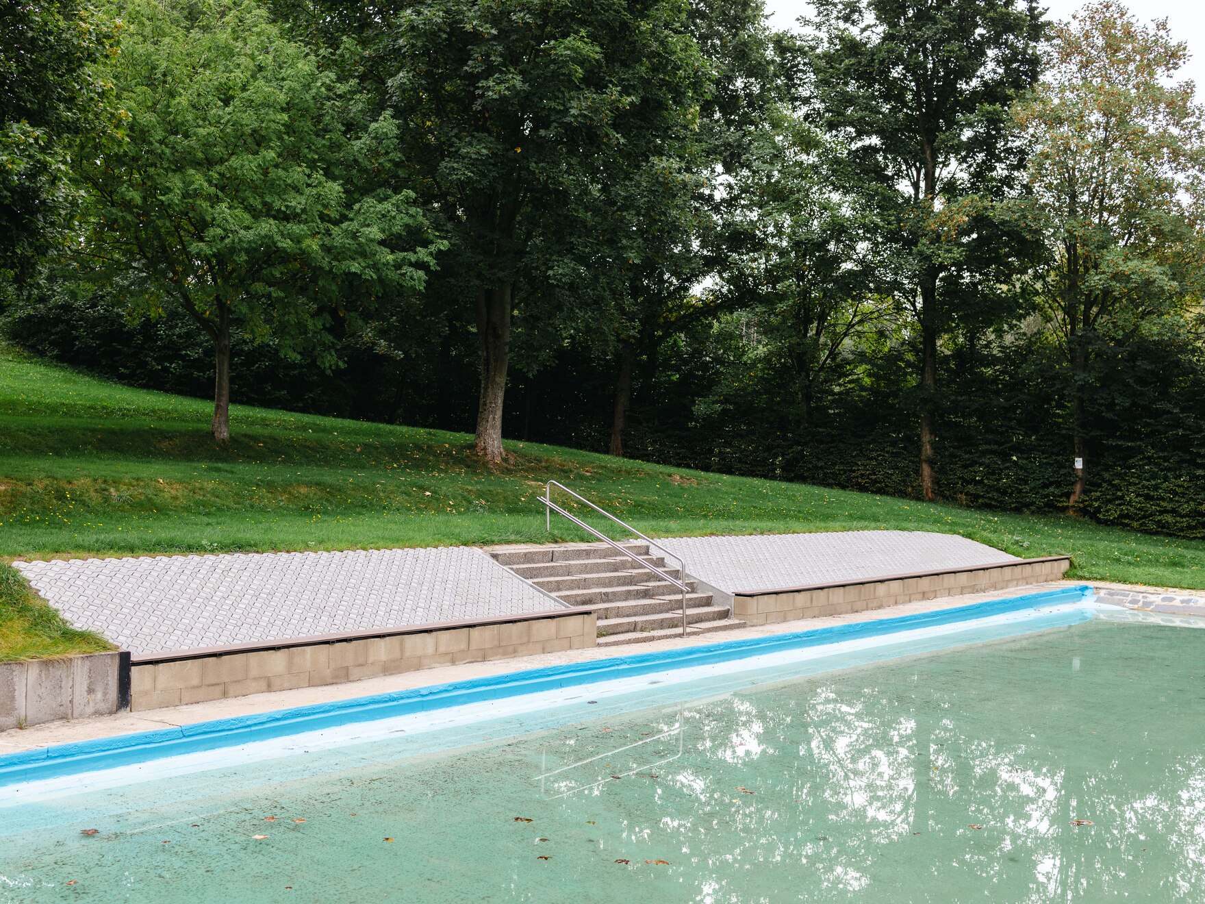 Blick auf ein Becken im Freibad Kottengrün. Eine Treppe führt ins Wasser. Dahinter die Liegewiese mit Bäumen und Büschen am Rand.