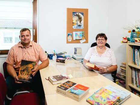 Zwei Personen sitzen in der Bücherei in Elstertrebnitz an einem Tisch, auf dem Bücher liegen, und halten selbst jeweils ein Buch in der Hand.