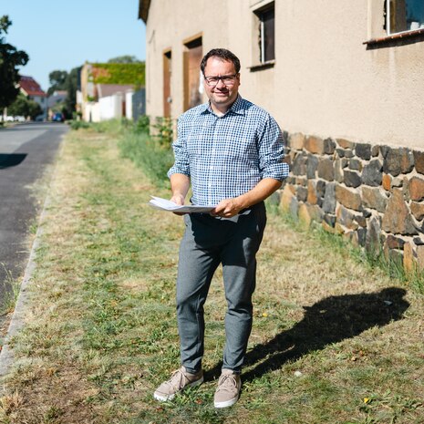 Ein Mitarbeiter der Stadt Taucha steht mit dem Dorfentwicklungskonzept in der Hand im Ortskern von Pönitz.