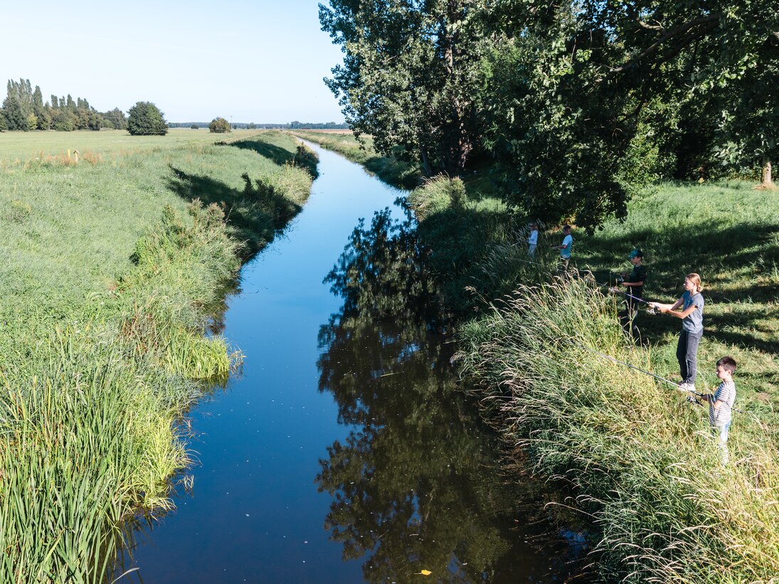 An einem Flusslauf im Grünen stehen Kinder und Jugendliche am Ufer und angeln.