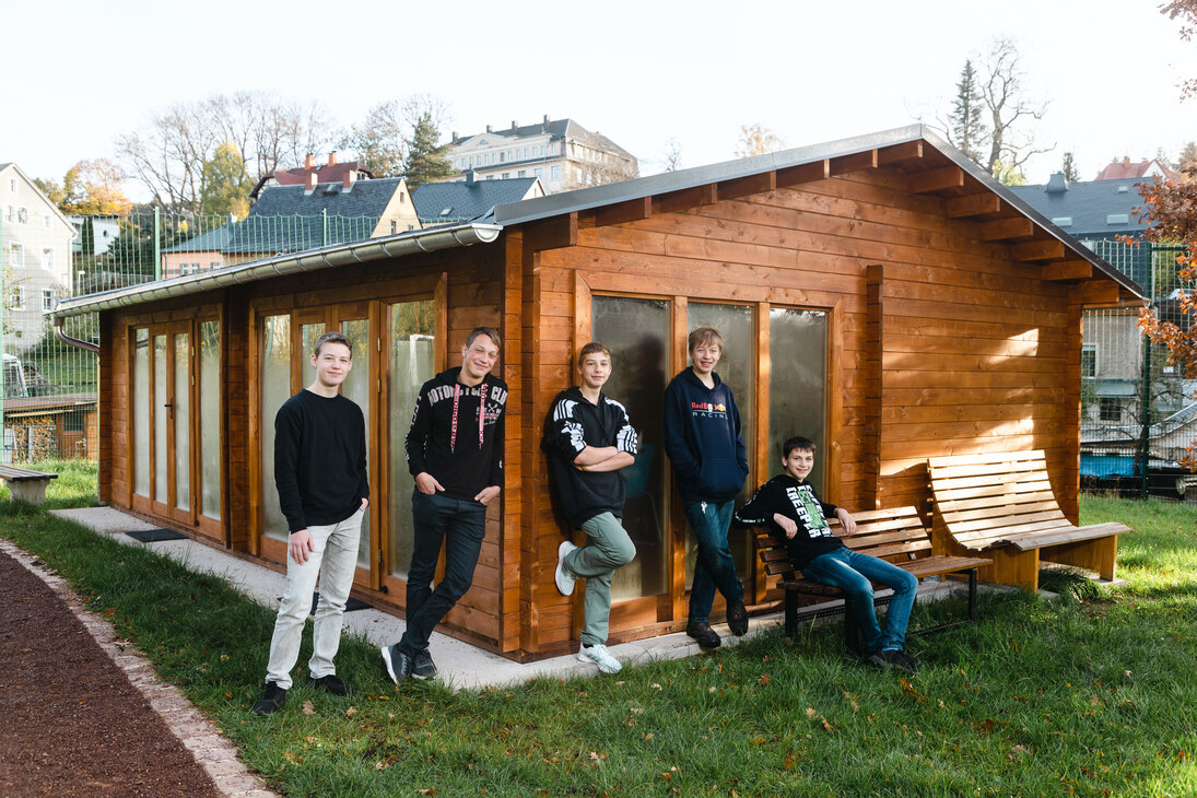 Fünf Jugendliche lehnen an einem Blockbohlenhaus auf dem Außengelände der Freien Evangelischen Oberschule in Burkhardtsdorf.