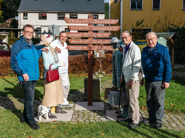 In einem grünen Park stehen vier Menschen neben zwei lebensgroßen Statuen in traditioneller Kleidung, begleitet von einem Hund. Ein dekorativer Wegweiser zeigt fantasievolle Ortsnamen mit Entfernungen in Kilometern.