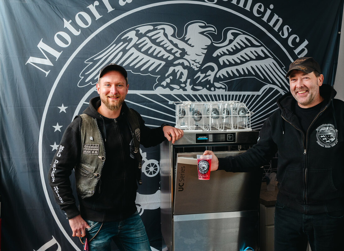Zwei junge Männer stehen vor einer Wand mit dem Vereinslogo. In der Mitte steht ein Geschirrspüler, darauf einige Mehrwegbecher. Ein Mann hält einen Becher in der Hand.