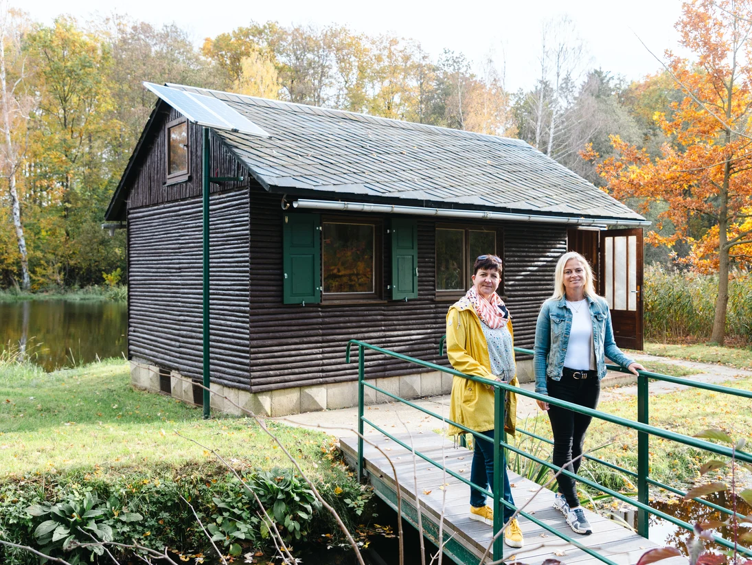 Zwei Frauen stehen auf einer kleinen Brücke. Diese führt auf die Insel mit dem umgestalteten Bungalow.