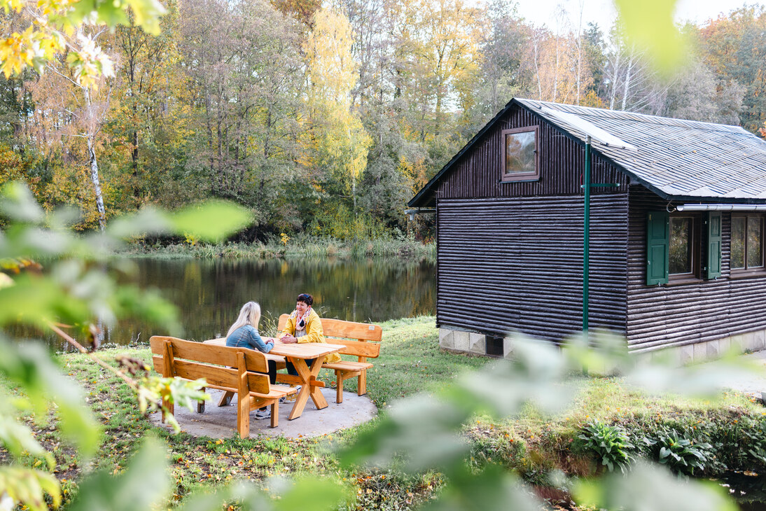 rechts im Bild ein Holzbungalow vor einem Teich. links eine Sitzgruppe, auf der 2 Personen Platz genommen haben
