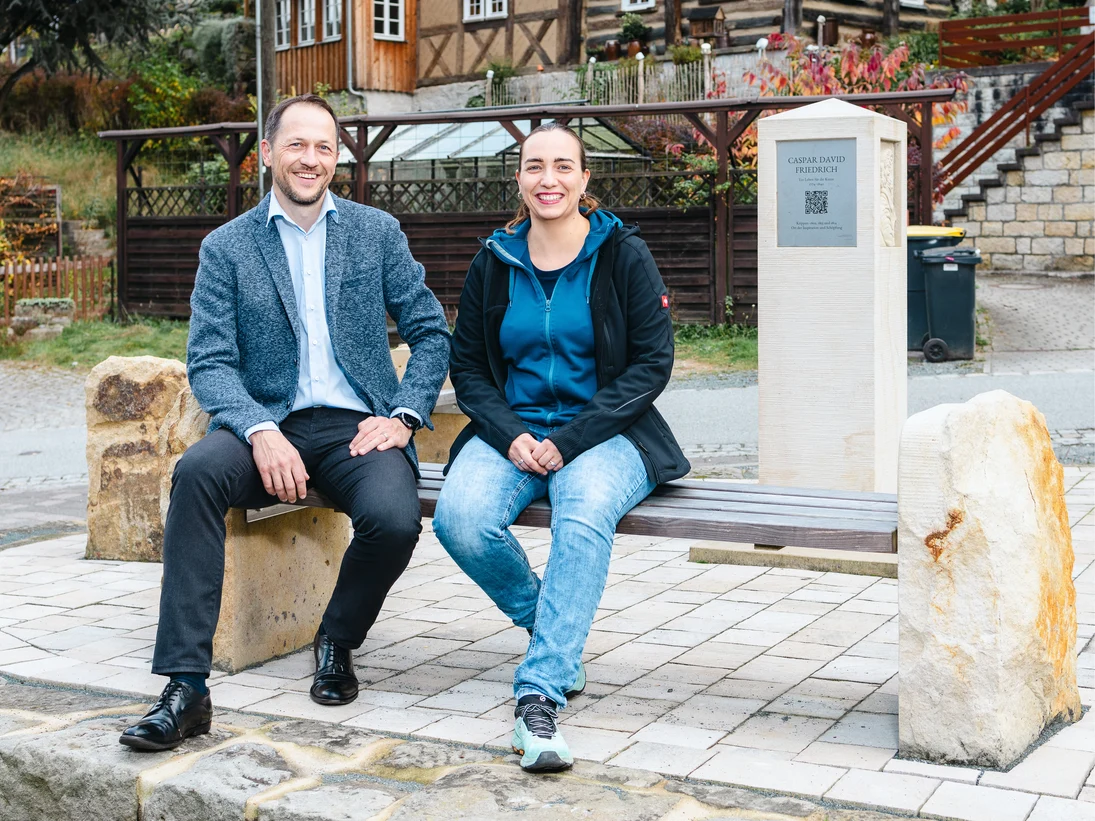 Zwei Menschen sitzen auf einer Bank. Daneben ist die neue Casper- David-Friedrich Stele zu sehen.
