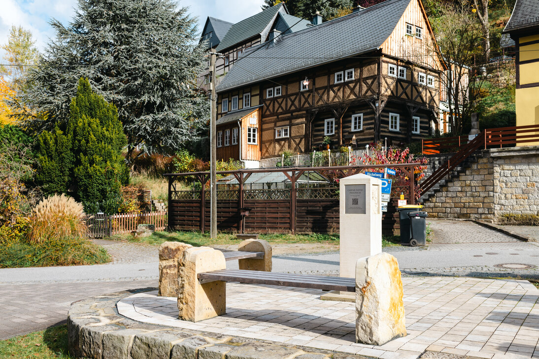 Zu sehen sind zwei Bänke, welche die Holzsitzfläche links und rechts mit Sandsteinblöcken hält. Im Hintergrund steht eine Stele mit einer Gedenkaufschrift
