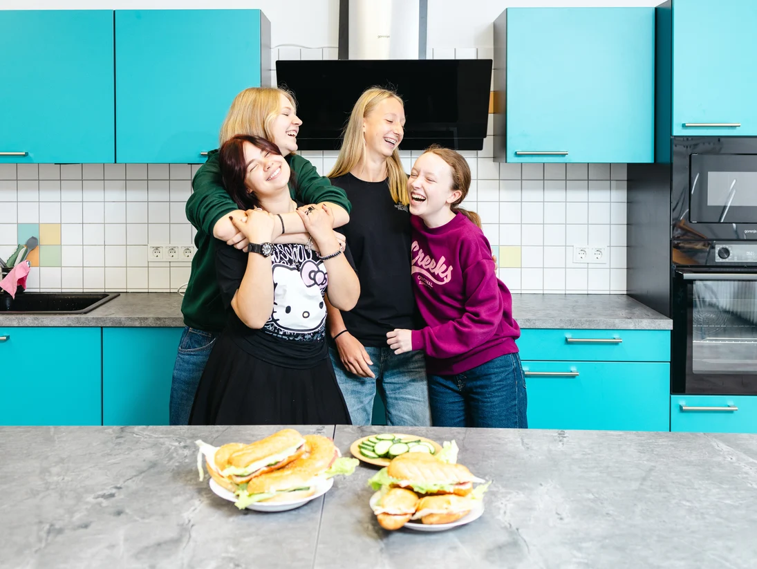 Vier Mädchen halten sich lachend in den Armen. Sie stehen mit frisch geschmierten Brötchen in der neuen Schulküche.