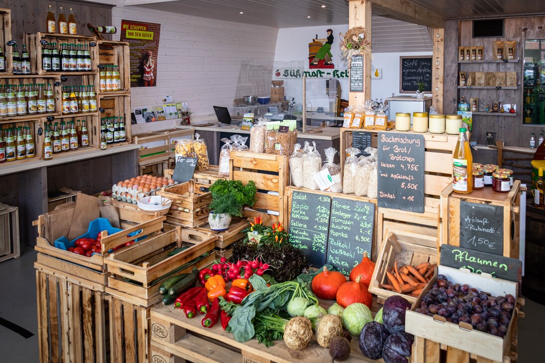 Ein Verkaufsstand mit regionalen Produkten, wie Obst, Gemüse, Eiern und Honig im Regionalladen. Regal im Hintergrund mit verschiedenen Saftsorten der Kelterei.