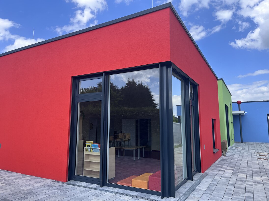 Blick auf das neue Gebäude der Kita Pockau-Lengefeld. Im Vordergrund ein großes, bis zum Boden reichendes Fenster. Die Fassade ist rot. Im Hintergrund ist die Fassade des Gebäudes grün und blau.