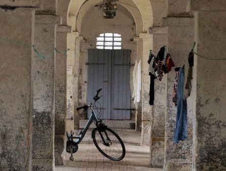 Blick zur Eingangstür des einstigen Kuhstalls. An den Säulen hängen Leinen mit Wäsche. An einer Säule lehnt ein Fahrrad.