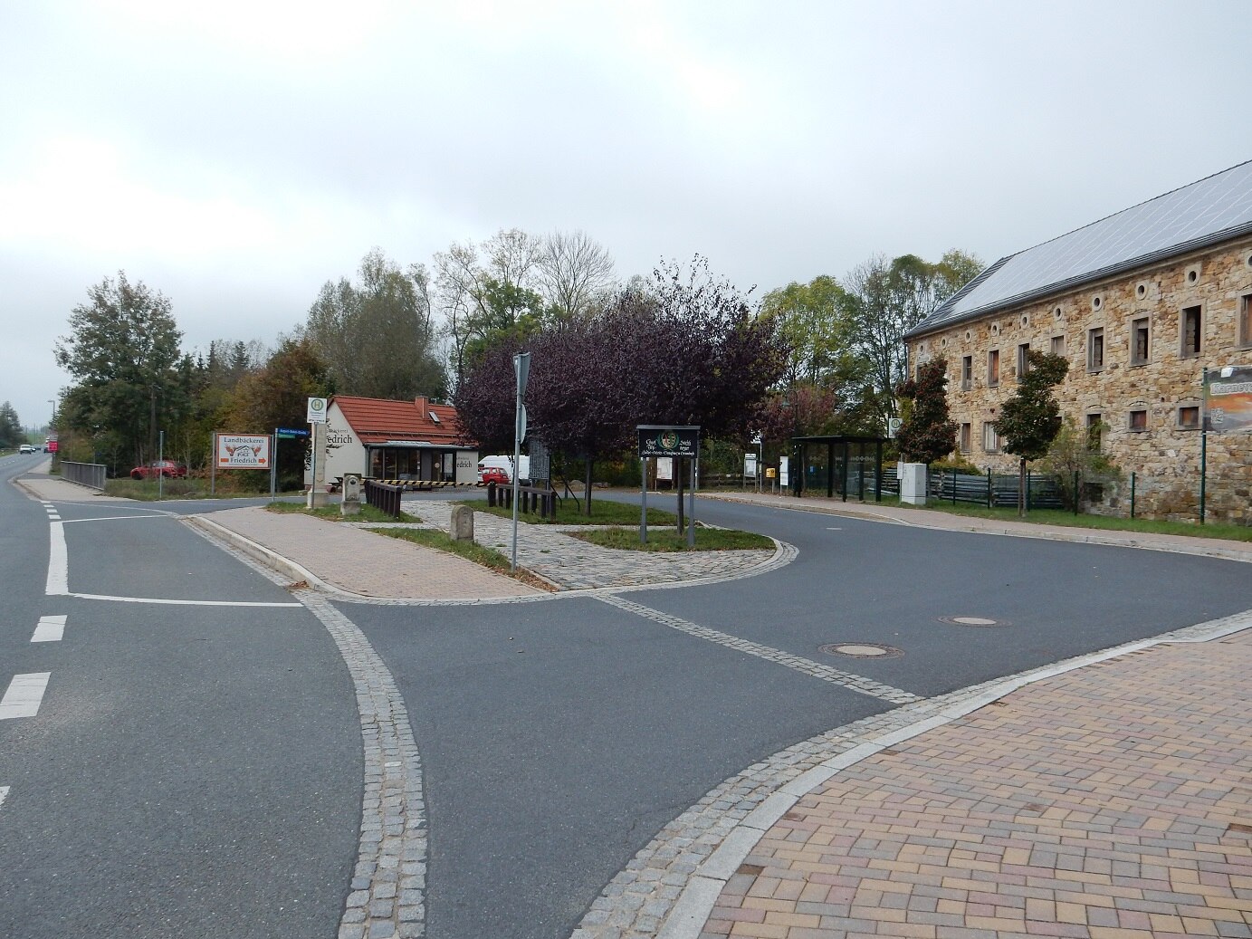 Umgestalteter Dorfplatz mit Straßen, historischer Anlage in der Mitte, Bushaltstelle, Fußweg und Bäckerei im Hintergrund
