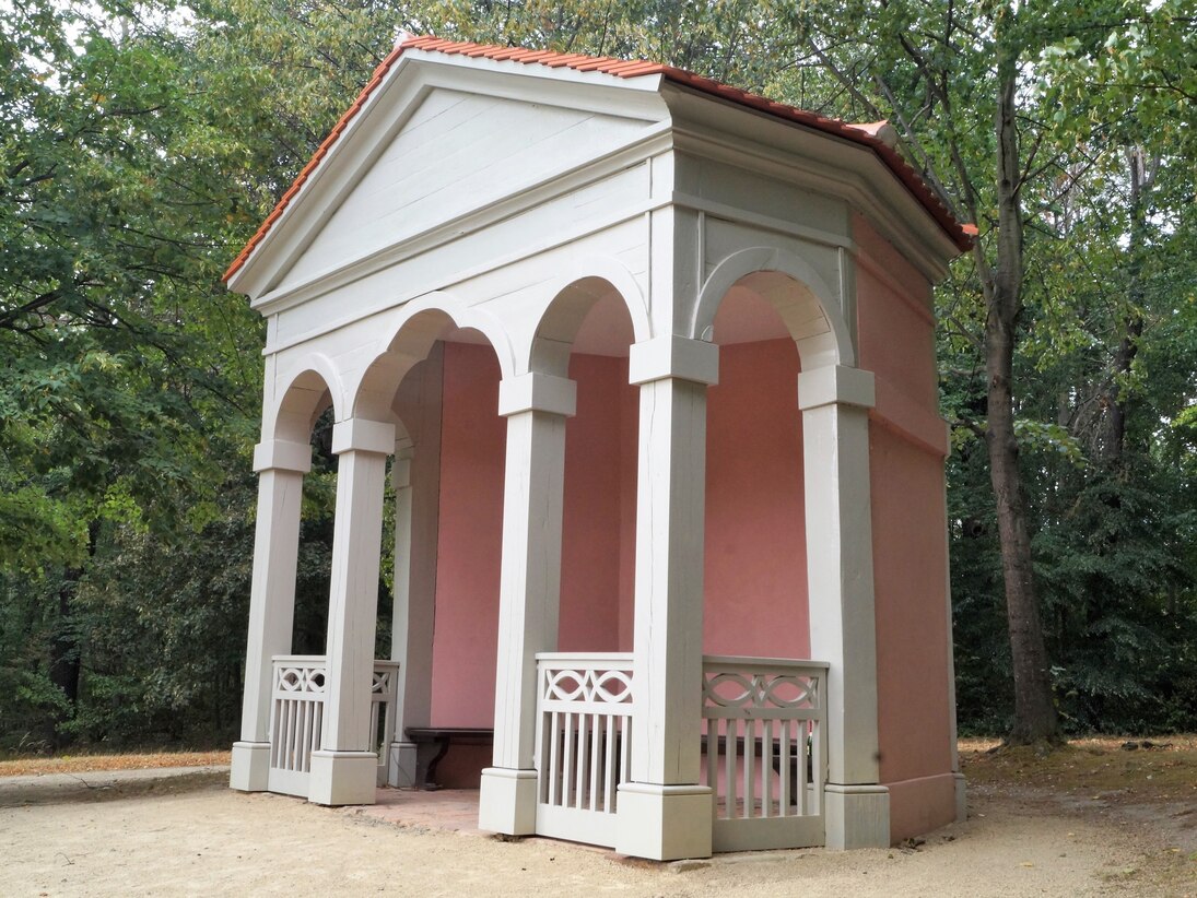 Sanierter Tempel mit weißen Säulen und englischroten Seitenwänden. Im Hintergrund stehen Rotbuchen. 