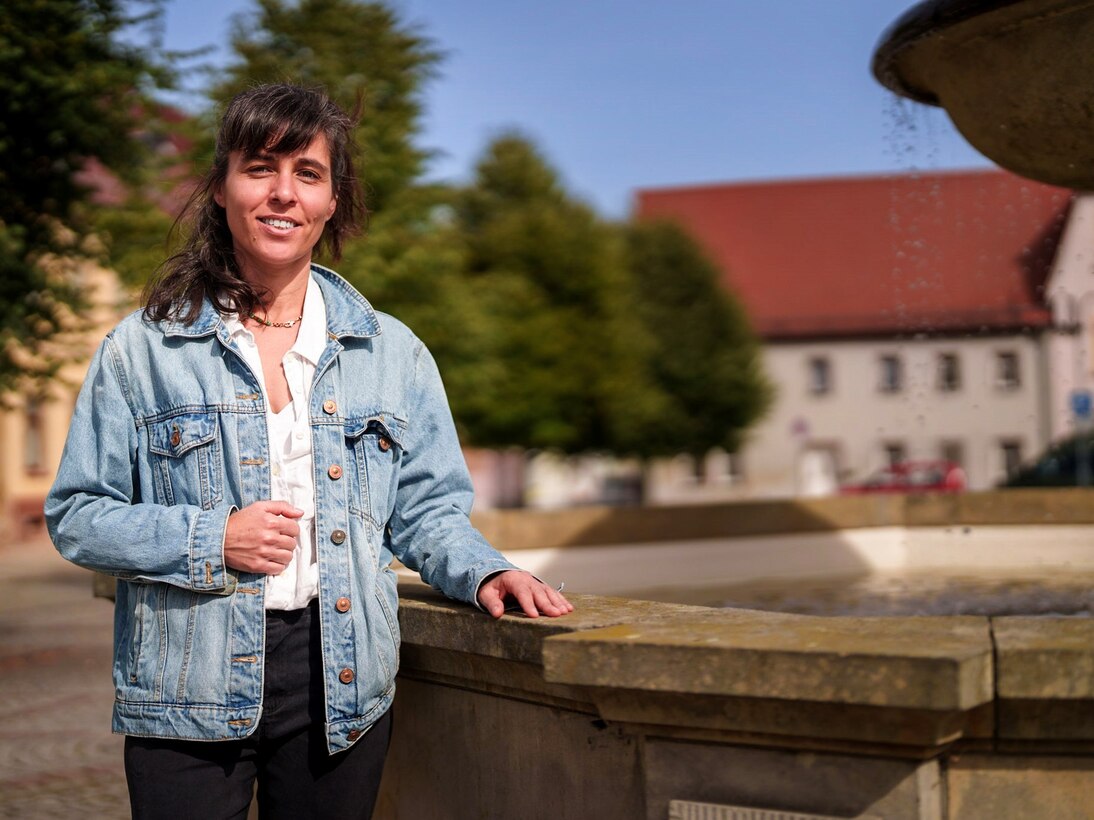 Die Künsterlin Michal Fuchs steht an einem Brunnen. Im Hintergrund ein Platz, Gebäude und Bäume.