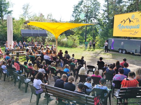 Auf einer Freilichtbühne am Waldrand steht eine große Leinwand mit dem Logo des Filmfestivals Eibenstock darauf. Davor sitzen viele Erwachsene und Kinder auf aufgestellten Bänken und Stühlen.