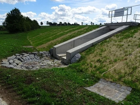 Das Bild zeigt eine Regenwasser-Überlaufstruktur in einem begrünten Hang. Ein betonierter Kanal leitet überschüssiges Wasser zu zwei mit Gittern abgedeckten Abflüssen, die von Steinen umgeben sind und der Erosionskontrolle dienen.