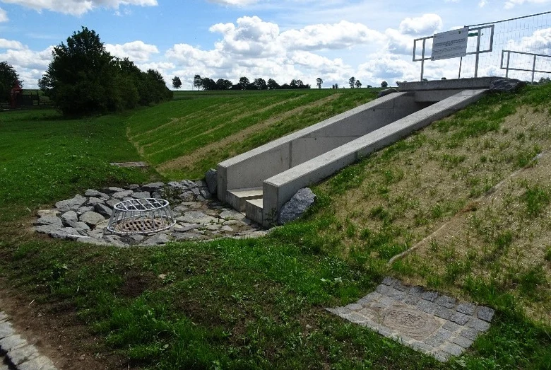 Das Bild zeigt eine Regenwasser-Überlaufstruktur in einem begrünten Hang. Ein betonierter Kanal leitet überschüssiges Wasser zu zwei mit Gittern abgedeckten Abflüssen, die von Steinen umgeben sind und der Erosionskontrolle dienen