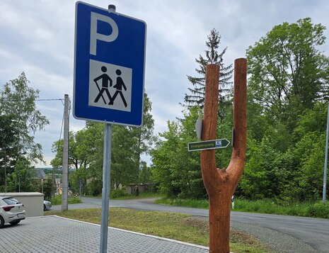 Wanderparkplatz mit blauem Parkplatzschild und hölzernem Wegweiser in Form einer Gabel mit grünem Schild 'Wernitzgrün' vor bewaldeter Umgebung.