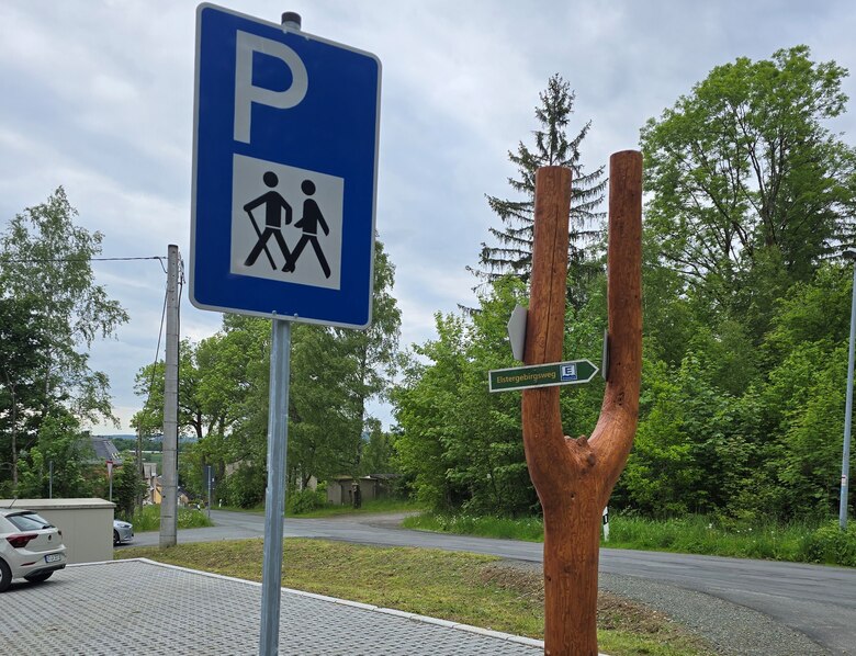 Wanderparkplatz mit blauem Parkplatzschild und hölzernem Wegweiser in Form einer Gabel mit grünem Schild 'Wernitzgrün' vor bewaldeter Umgebung
