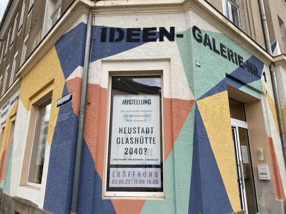 Die Ecke eines Gebäudes ist mit einem bunten geometrischen Wandbild in Blau, Gelb, Oran-ge und Beige gestaltet. An der Fassade befindet sich die Aufschrift „IDEEN-GALERIE 47“ sowie ein Plakat, das auf eine Kunstausstellung hinweist.